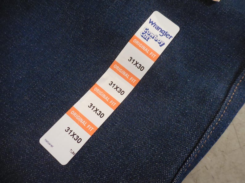 May include: A close-up of a blue denim fabric with a white tag that reads "Wrangler Cowboy Cut Original Fit" and lists the size "31X30" four times.