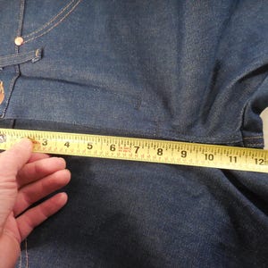 May include: A close-up of a blue denim fabric with a yellow measuring tape across it. The tape reads "3 4 5 6 7 8 9 10 11 12 13 14".