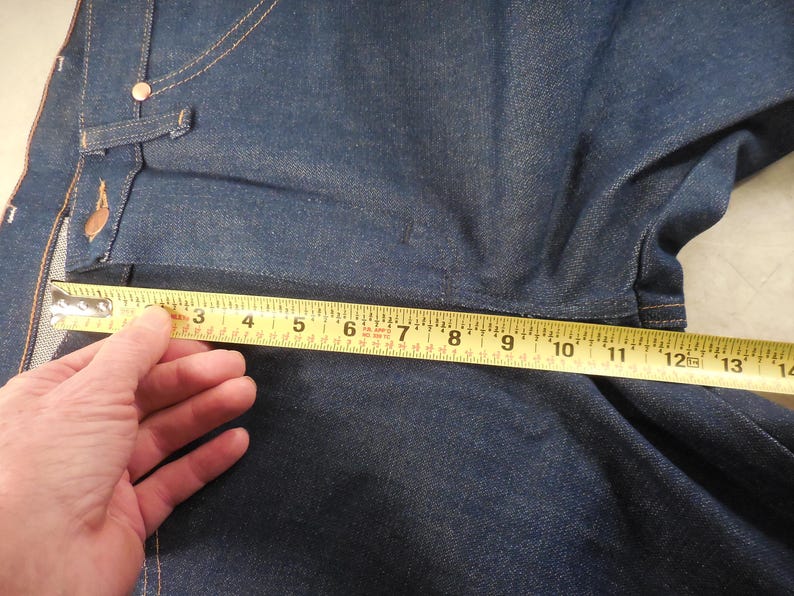 May include: A close-up of a blue denim fabric with a yellow measuring tape across it. The tape reads "3 4 5 6 7 8 9 10 11 12 13 14".