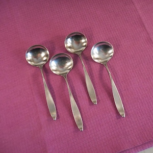 May include: Four shiny, silver soup spoons with rounded bowls and slender handles are arranged on a textured, magenta surface. The spoons are reflective, capturing the light and creating highlights.