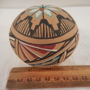 May include: A round, hand-painted ceramic vessel with a tan base color. The vessel features geometric designs in black, red, white, and turquoise. The top has a floral design. A wooden ruler is in the foreground.