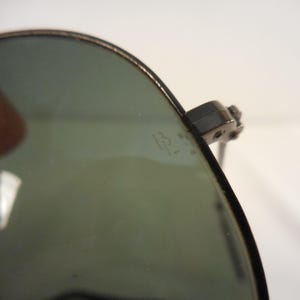 May include: Close-up of aviator sunglasses with a dark frame and green-tinted lenses. The sunglasses have a classic design, with a metal frame and a subtle logo etched on the lens. The lenses are slightly reflective.