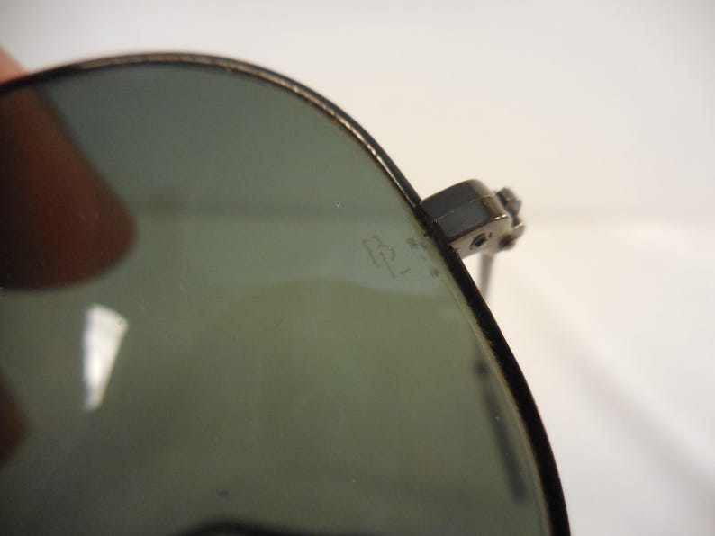 May include: Close-up of aviator sunglasses with a dark frame and green-tinted lenses. The sunglasses have a classic design, with a metal frame and a subtle logo etched on the lens. The lenses are slightly reflective.