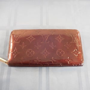 May include: A brown, zip-around wallet with a glossy finish. The wallet features a repeating pattern of embossed floral and geometric shapes, with a prominent logo in the center. The zipper pull is gold-toned.