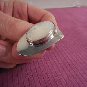 May include: A silver ring with an oval, off-white stone set on a rectangular base. The ring's design features a polished silver band and a raised setting for the stone. The ring is held against a purple background.