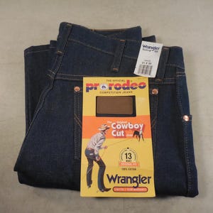 May include: A pair of blue denim jeans with a brown and yellow label that says "The Official ProRodeo Competition Jeans" and "Original Cowboy Cut". The label also has a picture of a cowboy riding a horse. The jeans have a Wrangler brand label on the back pocket.