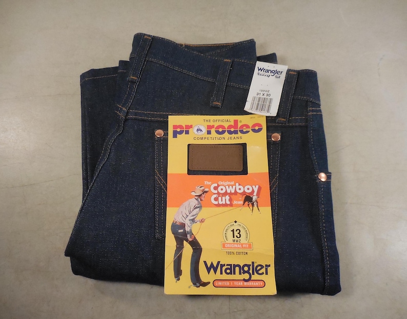 May include: A pair of blue denim jeans with a brown and yellow label that says "The Official ProRodeo Competition Jeans" and "Original Cowboy Cut". The label also has a picture of a cowboy riding a horse. The jeans have a Wrangler brand label on the back pocket.
