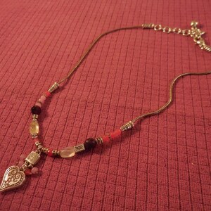 May include: A silver-toned necklace with a heart-shaped pendant and a variety of beads in shades of pink, red, and yellow. The pendant has an ornate design. The necklace is on a red textured surface.