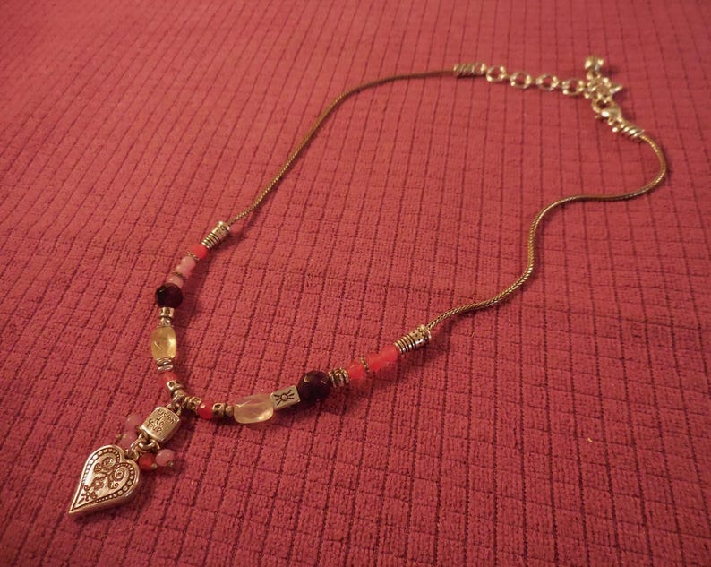 May include: A silver-toned necklace with a heart-shaped pendant and a variety of beads in shades of pink, red, and yellow. The pendant has an ornate design. The necklace is on a red textured surface.