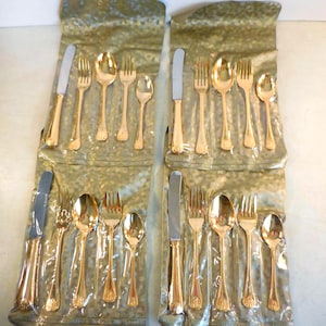May include: A set of gold-colored flatware, including knives, forks, and spoons, packaged in individual plastic sleeves. The silverware has a decorative design on the handles. The set appears to be for a formal dining setting.