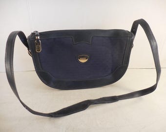 Puede incluir: Un bolso de hombro azul marino con una correa de cuero negro. El bolso tiene forma curva con ribete de cuero negro y un emblema dorado. El bolso tiene una cremallera dorada y un emblema dorado con el nombre de la marca.