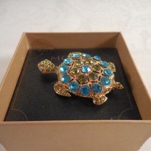 May include: A gold-toned turtle brooch featuring a shell encrusted with alternating bright blue and lime green faceted rhinestones. The head and flippers are decorated with smaller lime green stones. The brooch is presented in a square box.