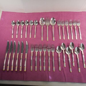 May include: A complete stainless steel flatware set with gold-colored accents. The set includes knives, forks, and spoons of various sizes, arranged on a magenta surface. The silverware has a shiny, reflective finish.