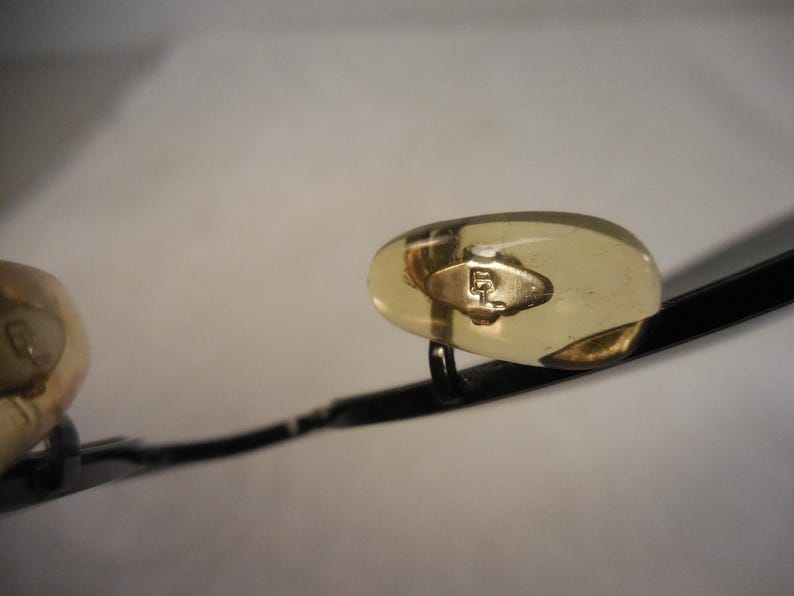 May include: Close-up of eyeglasses with a black frame and gold-colored nose pads. The nose pads have a gold-colored emblem. The lenses are not visible in the image.