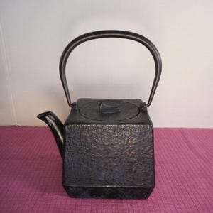 May include: Black cast iron teapot with a textured surface and a curved handle. The teapot has a square body, a spout, and a lid. This teapot is ideal for brewing tea.