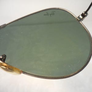 May include: Close-up of aviator sunglasses with green lenses and a silver frame. The Ray-Ban logo is visible on the lens. The nose pads are a light brown color. The sunglasses are set against a white background.