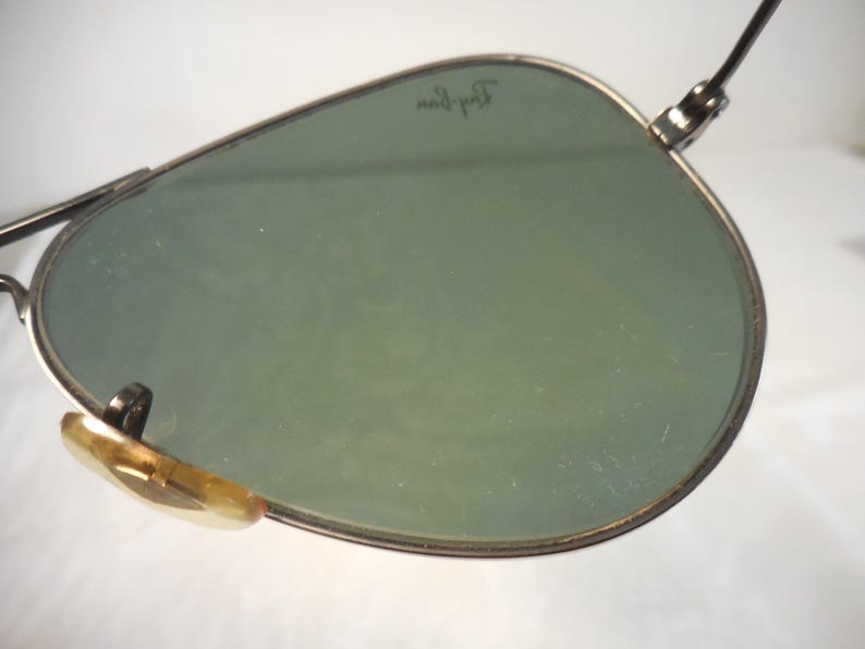 May include: Close-up of aviator sunglasses with green lenses and a silver frame. The Ray-Ban logo is visible on the lens. The nose pads are a light brown color. The sunglasses are set against a white background.