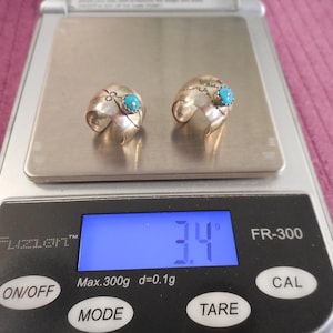 May include: Two silver-toned open-band rings, each featuring a small, round, bright blue stone set in the center. The rings are resting on a digital scale that displays a weight of 3.4 grams. The scale is labeled Fuzion FR-300.