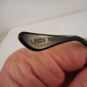 May include: Close-up of a black eyeglass arm. The text "L 2823 YNAS" is printed in silver on the inner surface of the arm. The arm is curved and has a glossy finish. The background is a blurred, neutral color.