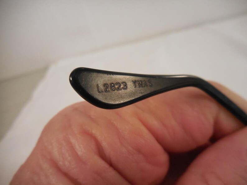 May include: Close-up of a black eyeglass arm. The text "L 2823 YNAS" is printed in silver on the inner surface of the arm. The arm is curved and has a glossy finish. The background is a blurred, neutral color.