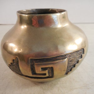 May include: A small, gold-colored ceramic vase with a geometric design. The vase has a rounded body and a slightly flared lip.