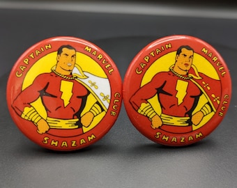 May include: Two red and yellow Captain Marvel Club buttons featuring a cartoon image of Captain Marvel in his red and yellow costume. The text on the buttons reads "Captain Marvel Club Shazam".