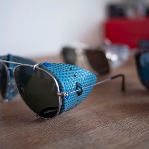 May include: Silver aviator sunglasses with a blue leather brow bar. The sunglasses have dark lenses and a silver frame.