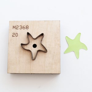 Peut inclure: Un gabarit de découpe en bois avec le numéro "20" et le code "M2368" imprimés dessus. Le gabarit est en forme d'étoile à cinq branches avec un trou au centre. Une découpe en papier vert d'une étoile à cinq branches est représentée à droite du gabarit.