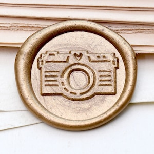 May include: A gold-colored wax seal with a camera design, featuring a heart above the lens. The seal is circular and sits on a stack of aged paper and a white surface. The camera design is detailed.