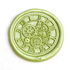 Può includere: Un sigillo di cera rotondo, verde chiaro, con un design a quadrante d'orologio. Il sigillo presenta numeri romani, ingranaggi e decorazioni. La cera ha una sottile lucentezza.