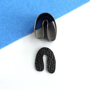 Puede incluir: Una herramienta de artesanía en cuero con forma de herradura de metal junto a una pieza de cuero recortada. La herramienta es plateada y el cuero es negro. El fondo es blanco con un cuadrado de fieltro azul en la esquina superior izquierda.