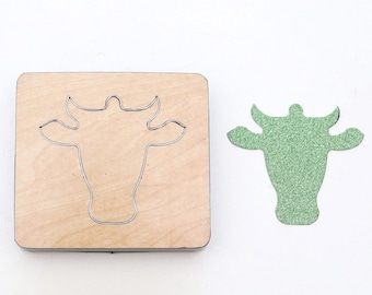 May include: A wooden die-cut with a cow head design. A green glitter cow head is cut out using the die.