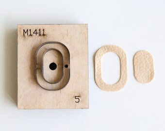 May include: A close-up of a leather craft punch with the number "M1411" and the number "5" printed on it. The punch is used to create oval-shaped holes in leather. Two pieces of leather have been punched with the tool, showing the oval-shaped holes.