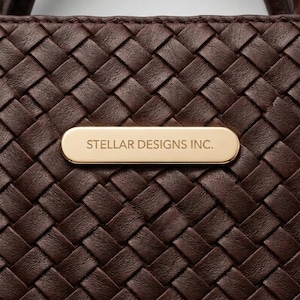May include: A dark brown woven leather handbag featuring a smooth, horizontal, gold-toned metal plaque centered on the front. The plaque is engraved with the text STELLAR DESIGNS INC. in a clean, sans-serif font. A rounded handle is visible at the top.