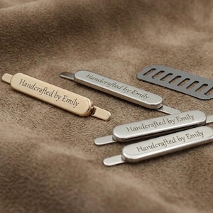 Könnte beinhalten: Eine Sammlung kleiner, länglicher Metall-Kleidungsetiketten in Gold- und Silbertönen, jeweils graviert mit dem Text Handcrafted by Emily. Die Etiketten haben dünne Befestigungsstifte an den Enden und ein dunkelgraues, rechteckiges Teil mit Schlitzen.