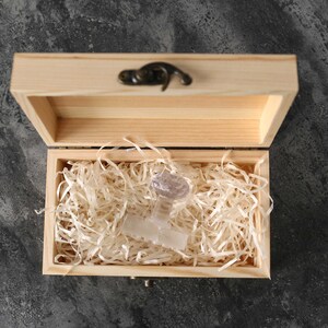 May include: A clear glass object with a unique design sits inside a wooden box lined with white shredded paper. The box has a hinged lid and a metal latch.