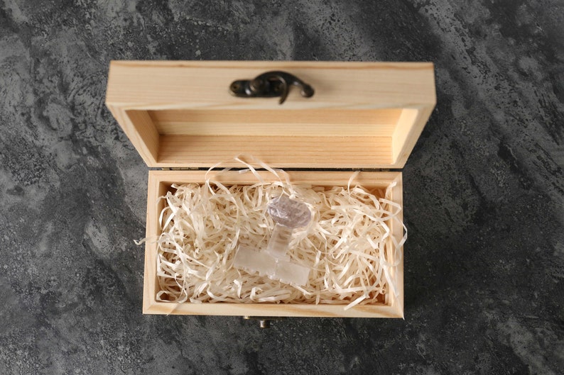 May include: A clear glass object with a unique design sits inside a wooden box lined with white shredded paper. The box has a hinged lid and a metal latch.