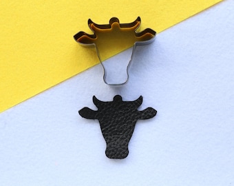May include: A metal cookie cutter in the shape of a cow's head, on a yellow and white background.