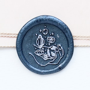 Puede incluir: Un sello de cera azul oscuro con un diseño blanco de una mano sosteniendo un planeta con estrellas y flores alrededor.