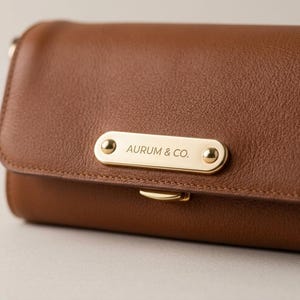 Puede incluir: Una cartera de cuero marrón con una placa metálica rectangular de tono dorado en la solapa frontal. La placa está asegurada con dos remaches dorados y muestra el texto grabado AURUM & CO. Un pequeño cierre metálico dorado es visible bajo la placa.