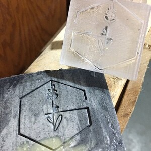May include: Two hexagonal rubber stamps, one clear and one black, with a floral design. The design features a stylized flower with a stem and leaves.