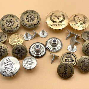 Puede incluir: Una colección de botones metálicos con temática náutica. Los botones presentan brújulas, veleros, faros y cascos de buceo. Los colores incluyen latón antiguo, plata y oro, con textos como "Naval Expedition" y "Oceanic Sport".