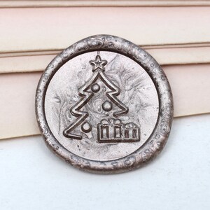 Puede incluir: Un sello de cera de color plateado con un diseño de árbol de Navidad y regalos.