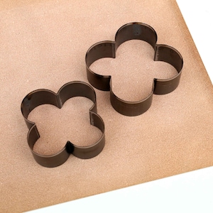Puede incluir: Dos cortadores de galletas de metal con forma de tr&eacute;bol de cuatro hojas. Los cortadores est&aacute;n sobre una superficie marr&oacute;n.