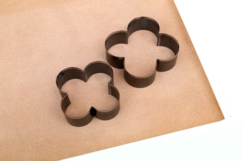 Puede incluir: Dos cortadores de galletas de metal con forma de tr&eacute;bol de cuatro hojas. Los cortadores est&aacute;n sobre una superficie marr&oacute;n.