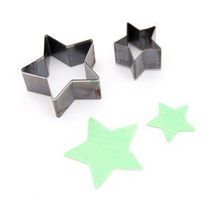 May include: Three metal star-shaped cookie cutters, two are silver and one is a lighter shade of silver. Two green felt stars are also shown.