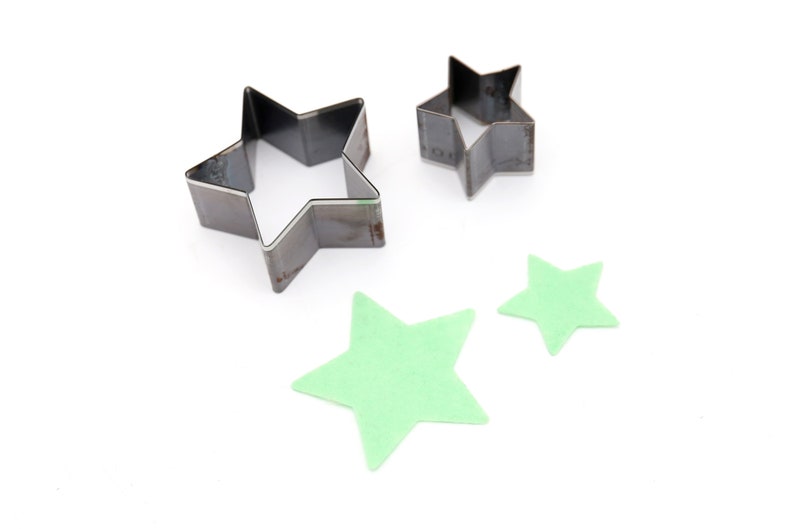 May include: Three metal star-shaped cookie cutters, two are silver and one is a lighter shade of silver. Two green felt stars are also shown.