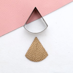 May include: A metal template in the shape of a teardrop is placed on top of a gold leather teardrop shape.