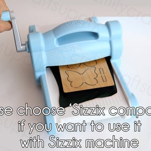 May include: A light blue die-cutting machine with a hand turning the crank. The machine is cutting out a butterfly shape from a piece of wood. The text on the machine reads "Sizzix". The text below the machine reads "Please choose 'Sizzix compatible' if you want to use it with Sizzix machine."