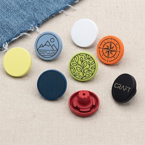 Könnte beinhalten: Eine Sammlung bunter Jeansknöpfe in verschiedenen Designs und Farben. Die Knöpfe umfassen eine Berglandschaft, einen Kompass und ein Blattdesign. Ein Knopf ist schwarz mit dem Wort "CRAFT". Weitere Knöpfe sind gelb, weiß und blau.